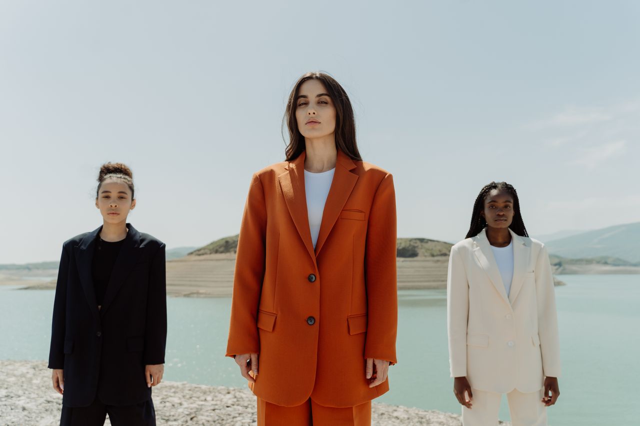heros-img Three stylish women in suits posing confidently outdoors by a serene lake.